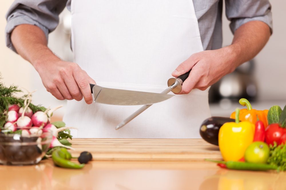 Cook sharpening knives with food and vegetables on the kitchen counter. 