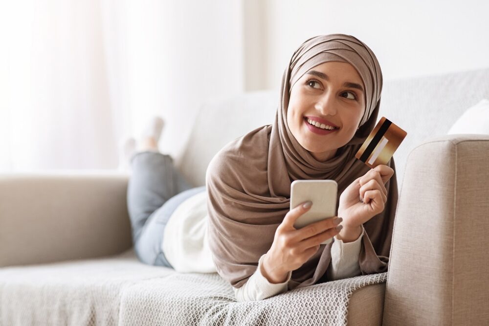 Online shopping. Pensive girl in hijab with smartphone and credit card lying on couch, thinking about internet purchases or food delivery
