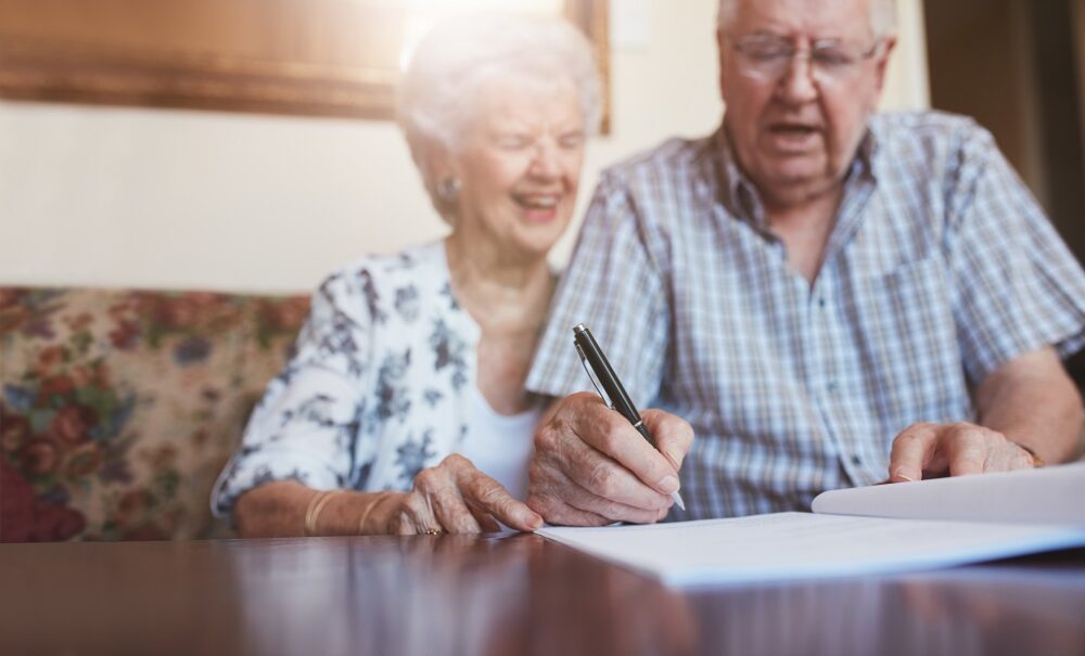 Portrait of a senior couple signing documents. Senior caucasian man and woman sitting on sofa and signing some paperwork, focus on hands.